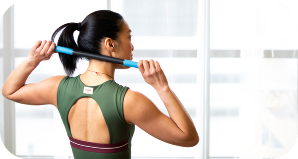Woman in green sportswear using a RAD ROD | RAD wellness recovery tool to massage her upper back indoors by a large window.