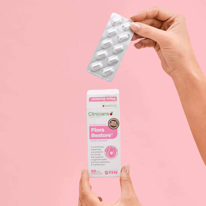 Hand holding a packet of 'Clinicians Flora Restore' capsules against a pink background.