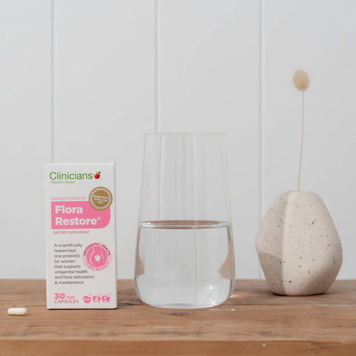 Clinicians Flora Restore supplement box with a glass and decorative item on a wooden surface.