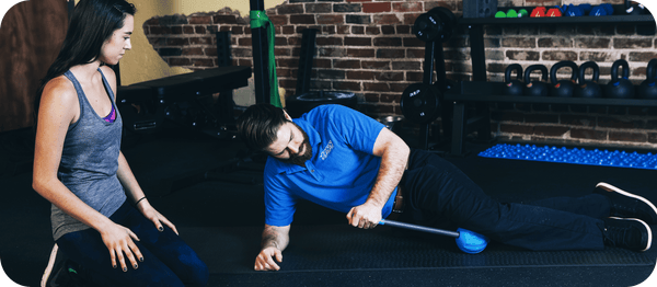 Man using RAD ATOM | RAD blue massage tool on side while a woman watches in gym setting, demonstrating recovery and wellness device for body care and muscle relief.